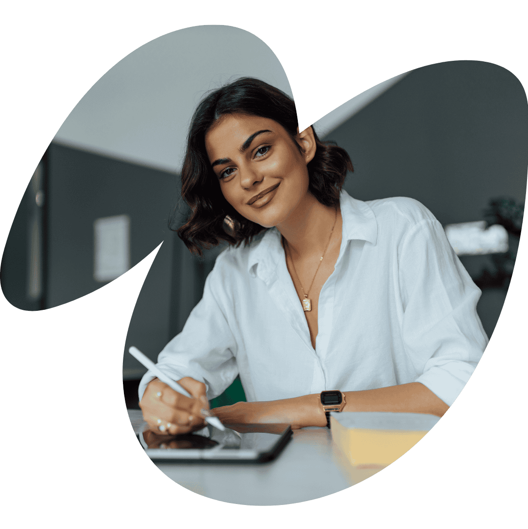 Femme brune en chemise blanche qui travaille sur une table, elle est souriante et suit un accompagnement individuel avec une coach